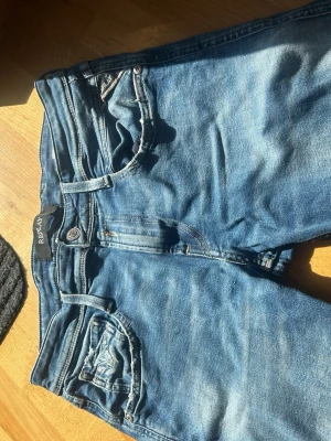 Blå slitna jeans från Replay - Säljer ett par blå jeans från Replay med snygga slitningar på ena benet. Jeansen har klassisk femficksmodell, dragkedjegylf och smal passform. Perfekta för en avslappnad och trendig look.