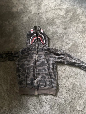 Camo hoodie med hajmotiv från BAPE - Grym camoflagemönstrad hoodie från BAPE med hajmotiv på huvan och broderade bokstäver och tiger på baksidan. Dragkedja hela vägen och två fickor fram. Perfekt för streetwear-stilen.