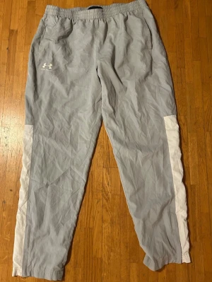 Grå byxor från Under Armour XL - Snygga grå träningsbyxor från Under Armour i storlek XL. Byxorna har vita paneler längs sidorna och elastisk midja med snörning. Tillverkade i lätt polyester med meshfoder för extra komfort. Perfekta för sport, friluftsliv och chill.