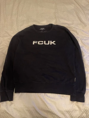 Svart sweatshirt från FCUK - Cool svart sweatshirt från FCUK med vit logga tryckt på bröstet. Tröjan har rund halsringning, ribbade muddar vid ärmslut och nederkant. Perfekt för en avslappnad och trendig stil.