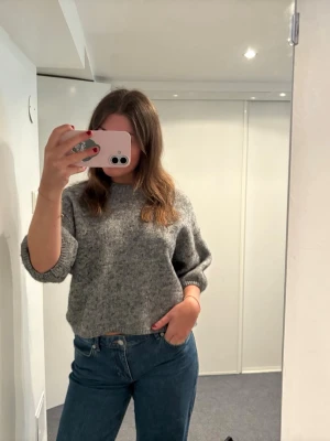 Grå stickad tröja med rund hals - Mysig grå stickad tröja med avslappnad passform och rund hals. Tröjan har långa ärmar och ribbade muddar vid ärmslut och nederkant. Perfekt att matcha med jeans för en enkel och stilren look.