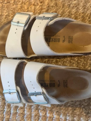 Vita Birkenstock sandaler med spännen - Klassiska vita sandaler från Birkenstock med två breda remmar och justerbara metallspännen. Fotbädden är formad för extra komfort och sulan är i kork. Perfekta för en avslappnad och trendig stil.