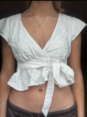 Vit omlottopp med knytband - Säljer en vit omlottopp med knytband i midjan och volang nedtill. Toppen har v-ringning och är ärmlös med breda axelpartier. Perfekt för sommardagar och enkel att styla till olika looks.