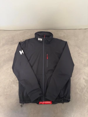 Helly Hansen vindjacka svart - Snygg svart vindjacka från Helly Hansen med HH-logga på kragen och ärmen. Jackan har hel dragkedja framtill, bröstficka med röd dragkedja och justerbar nederkant. Tillverkad i ett lätt och slitstarkt material som passar perfekt för blåsiga dagar. PRIS KAN DISKUTERAS