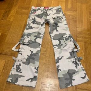 Camo cargobyxor med spännen - Säljer ett par  cargobyxor med grått camomönster. Byxorna har flera stora fickor, coola spännen längs benen och utsvängd passform. 