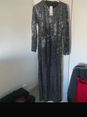 Glittrig långklänning med slits - Snygg långklänning med glittrigt mönster i svart och silver. Klänningen har långa ärmar, en markerad slits framtill och en kroppsnära passform. Perfekt för dig som vill sticka ut på festen! Materialet känns stretchigt och bekvämt.