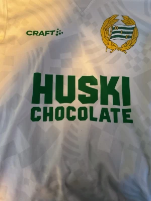 Hammarby vit matchtröja Craft - Säljer en vit Hammarby matchtröja från Craft med grönt tryck 'HUSKI CHOCOLATE' på bröstet och klubbmärke i gult och grönt. Tröjan har korta ärmar med grönt mönster vid ärmslut och V-ringad hals. Perfekt för fotbollsfans! 