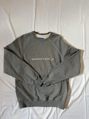 Grå sweatshirt från Jacob Cohën - Grå sweatshirt från Jacob Cohën med trycket 'remember to Smile' och en liten blå/vit detalj på bröstet. Tröjan har rund halsringning, raglanärmar och ribbade muddar vid ärmslut och nederkant. Perfekt för en avslappnad och stilren look.