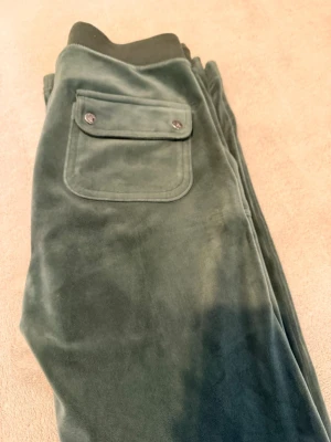 Mörkgröna velourbyxor med fickor - Notera att på bild 2 ser du på bandet att de fattas en metalbit. Mörkgröna mjukisbyxor i mjuk velour med bred resår och dragsko i midjan. Byxorna har stora fickor fram och en ficka bak med lock och knappar. Perfekta för chill dagar och har en snygg detalj med broderad logga på ena fickan.