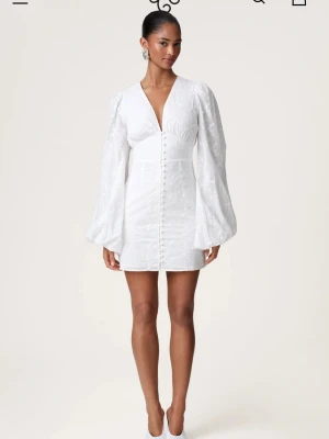 Vit kortklänning med puffärm och brodyr - Superfin vit kortklänning med v-ringning, puffiga långärmar och broderade blommor. Klänningen har knappar framtill och är gjord i ett lätt, luftigt material som passar perfekt till sommaren. Snygg och trendig modell med markerad midja och voluminösa ärmar. Använd 1 gång. Inga defekter.