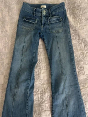 Blå bootcut jeans från NELLY - Säljer ett par ljusblå bootcut jeans från NELLY med hög midja och dekorativa sömmar längs benen. Jeansen har två stora fickor framtill och dubbla knappar i midjan. Perfekta för en avslappnad och trendig look.