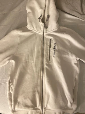 Vit hoodie från Sail Racing - Vit hoodie från Sail Racing med dragkedja storlek 170/s/m. Märkets logga är broderad på bröstet. Hoodien har justerbar huva med snören och är tillverkad i mjukt bomullsmaterial. Perfekt för en clean och sportig look. Märket på luvan har blekts lite.