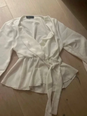 Vit omlottblus från Miss Selfridge - Stilren vit omlottblus från Miss Selfridge med knytband i midjan och v-ringad hals. Blusen har långa ärmar och ett lätt, mjukt tyg som ger en avslappnad men chic look. Perfekt att matcha med jeans eller kjol för en trendig outfit.