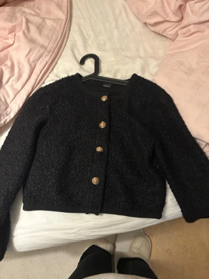 Svart bouclékofta med guldknappar - Stilren svart bouclékofta från gina tricot med rund halsringning och fyra dekorativa guldknappar framtill. Kortare modell med lång ärm och mjuk, fluffig textur. Perfekt för dig som gillar trendiga detaljer och en klassisk look.