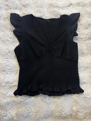 Svart topp med volangärmar  - Säljer en svart topp med v-ringning och volangdetaljer vid ärmar och nederkant. Toppen har smockad midja som ger en snygg passform och är tillverkad i ett stretchigt material. Perfekt att styla med jeans eller kjol.