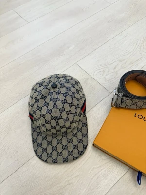 Gucci beige keps med GG-mönster - Säljer en beige keps från Gucci med klassiskt GG-monogram över hela kepsen och röda samt mörkblå ränder på sidorna. Justerbar rem bak i svart läder. Perfekt accessoar för dig som gillar lyxiga detaljer och streetstyle.