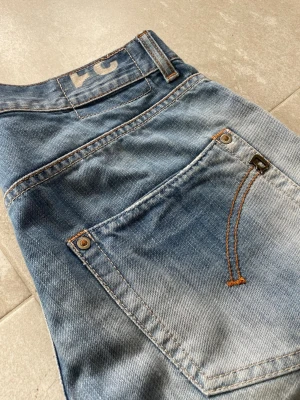 Blå slitna jeans från DonDup - Säljer ett par blå jeans från Dondup med snyggt slitna detaljer och klassisk femficksmodell. Jeansen har kontrastsömmar i orange och en rak passform. Materialet är mjuk denim i bomull som ger en avslappnad look. Mått 📏 Totallängd: 105cm Midjelängd: 40cm Innersömmlängd: 81cm Lårviddlängd: 27cm Skador 🔧: Litet hål vid innersömmen, små skador vid benöppningarna, visas på bild