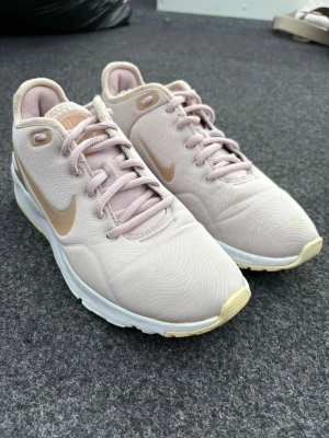 Ljusrosa Nike sneakers med beige detaljer - Snygga ljusrosa Nike sneakers med beige swoosh och detaljer. Ovandelen är i textil med subtilt mönster, och sulan är vit med beige inslag. Skorna har klassisk låg profil och matchande ljusrosa snören. Perfekta för en trendig och sportig look.