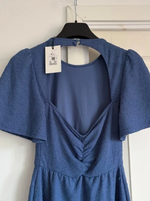 Blå kortklänning med v-ringning - Snygg blå kortklänning med strukturmönster, v-ringning framtill och öppen rygg med knapp i nacken. Klänningen har korta, vida ärmar och markerad midja för en feminin siluett. Perfekt för dig som vill ha en stilren men ändå unik look. Storlek Xs