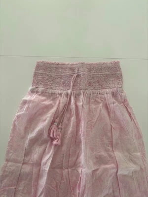 Ljusrosa långkjol med vitt mönster och tofsar  - Ljusrosa lång kjol från Gina Young med vita detaljer och två tofsar som har små vita pärlor på sig.