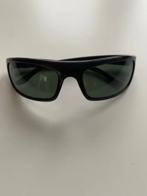 Ray-Ban solglasögon  - Solglasögon från Ray-Ban.Den här modellen är utsåld. Modellen har en omslutande bågform med svarta bågar och gröna glas.Det finns ingraverade RB på vänster lins,ett tecken på äkthet.