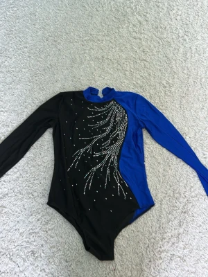 Blå och svart gymnastikdräkt - Säljer en långärmad gymnastikdräkt i svart och blått med glittrande stenar på framsidan. Dräkten har hög hals och är tillverkad i stretchigt material som passar perfekt för träning eller tävling inom gymnastik eller dans.