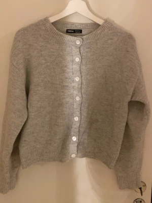 Ljusgrå kofta från SHEIN - Mysig ljusgrå kofta från SHEIN med rund halsringning och vita knappar framtill. Koftan har ribbade muddar vid ärmslut och nederkant, samt en mjuk och fluffig känsla. Perfekt att slänga på sig över en t-shirt eller linne.