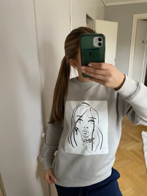 Grå/beige sweatshirt med tryck  - Grå/beige sweatshirt med ett stort tryck på framsidan och texten 'ONE DAY IN NEW YORK' på ryggen. Tröjan har rund hals, ribbade muddar och är gjord i mjukt material, perfekt för en avslappnad look. Endast använd vid 1-2 tillfällen.