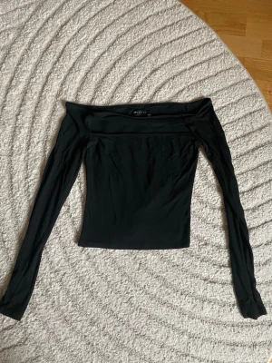 Svart offshoulder topp från Gina Tricot - Trendig svart offshoulder topp från Gina Tricot med lång ärm och tight passform. Tillverkad i mjuk och stretchig polyester som sitter snyggt på kroppen. Perfekt att styla med höga jeans eller kjol för en cool look.