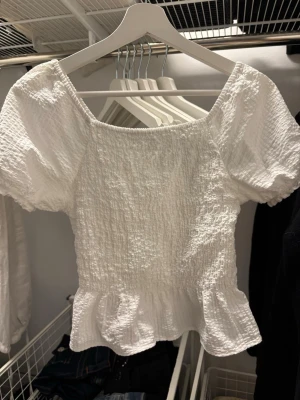 Vit smockad topp med volangkant - Söt vit topp med smockad struktur, korta puffärmar och volangkant nedtill. Toppen har en fyrkantig ringning fram och djupare rygg med knappar. Tillverkad i ett lätt, texturerat tyg som känns luftigt och somrigt. Sitter som XS ungefär💓