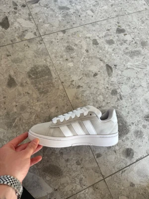 Vita Adidas sneakers med mockadetaljer - Snygga vita Adidas sneakers med klassiska tre ränder i vitt på sidan. Skon har en låg profil och är tillverkad i mocka och textil med vita skosnören och vit sula. Perfekt för dig som gillar stilrena och tidlösa sneakers.