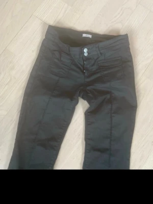 Nelly coated jeans  - Snygga svarta lackbyxor med bootcut-modell och dubbla silverfärgade knappar i midjan. Jeansen har framfickor och dekorativa sömmar. Perfekta för dig som gillar en klassisk men trendig look. Lite slitna längst ner 