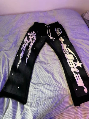 Svarta mjukisbyxor Hellstar print - Svarta mjukisbyxor från Hellstar med stora vita tryck längs ena benet och coola grafiska detaljer. Byxorna har dragsko i midjan och raka ben. Materialet är mjukt och bekvämt, perfekt för en avslappnad streetwear-look.