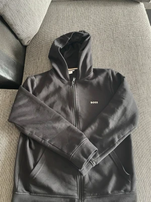 Svart zip hoodie från BOSS - Snygg svart hoodie från BOSS med dragkedja framtill och liten vit BOSS-logga på bröstet. Klassisk modell med huva och två fickor. Perfekt för en clean och stilren look. Tillverkad i mjukt bomullsmaterial som känns skönt mot huden.