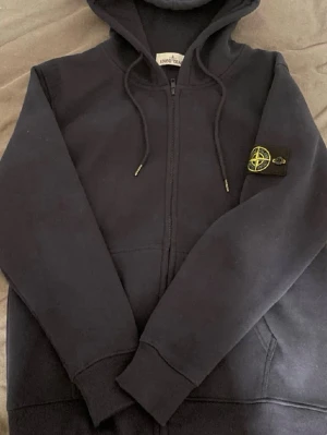 Mörkblå hoodie från Stone Island - Snygg mörkblå hoodie från Stone Island med dragkedja framtill och klassisk patch på ärmen. Hoodien har justerbar huva med snören och ribbade muddar. Perfekt för dig som gillar streetwear och vill ha en stilren look.