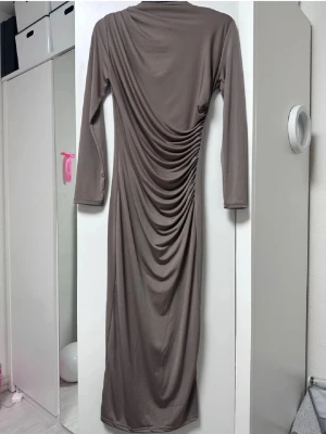 Lång brun klänning med drapering - Säljer en långklänning i mjukt, stretchigt material med snygg drapering längs sidan. Klänningen har lång ärm och en enkel, stilren design i en brun nyans. Perfekt för dig som gillar minimalistisk stil med en twist.