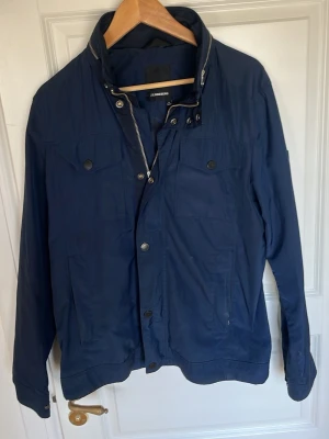 Marinblå vindjacka från J.Lindeberg - Säljer denna J Lindeberg jackan som är i ett använt skick vilket man kan se på första bilden där man ser lite fläckar. Men annars en jättesnygg jacka som funkar galant. Skriv vid funderingar!