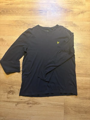 Mörkblå långärmad topp Lyle & Scott - Snygg mörkblå långärmad topp från Lyle & Scott med klassisk gul logga på bröstet. Enkel och stilren design med rund halsringning. Tröjan har ett litet hål vid vänster arm.