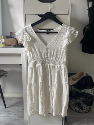 Vit spetsklänning med volangärmar - Söt vit klänning med spetsdetaljer och volangärmar. Klänningen har v-ringning, knappar framtill och smock i midjan för en bekväm passform. Perfekt för varma dagar och har en romantisk känsla med sina fina detaljer.