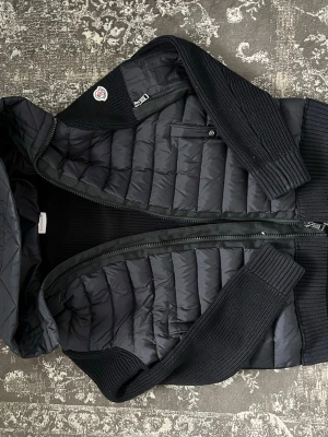 Svart Moncler cardiganjacka  - Passar den om du är mellan 174-184 Snygg svart cardiganjacka från Moncler med quiltad front och rygg i dun, stickade ärmar och sidor samt huva. Jackan har dragkedja framtill, bröstficka med knapp och Moncler-logga på ärmen. Perfekt för kalla dagar och riktigt trendig look.