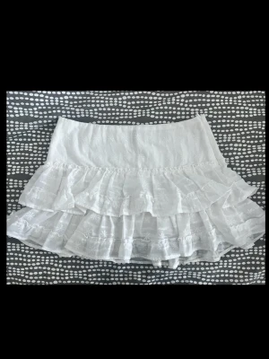 Vit volangkjol med broderi - Söt vit kjol med flera lager volanger och broderade blommor. Kjolen har en bred midja och är gjord i ett tunt, luftigt tyg som känns perfekt för sommaren. Detaljerade broderier ger kjolen en romantisk och trendig look.