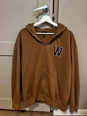 Brun hoodie med dragkedja från H&M - Säljer en brun hoodie från H&M i relaxed fit med dragkedja framtill och stor ficka på magen. På bröstet finns ett broderat W. Hoodien har huva och ribbade muddar vid ärmar och nederkant. Perfekt för en chill och avslappnad stil.