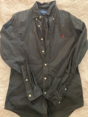 Svart skjorta från Ralph Lauren - Klassisk svart skjorta från Ralph Lauren med röd broderad logga på bröstet. Skjortan har långa ärmar, vita knappar och är tillverkad i mjuk bomull. Perfekt för en stilren och tidlös look.