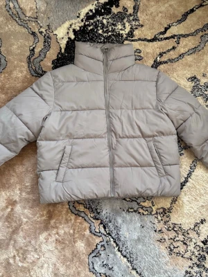 Beige pufferjacka med hög krage - Säljer en beige pufferjacka med hög krage och dragkedja framtill. Jackan har två snedställda fickor och quiltad design som ger en riktigt puffig look. Perfekt för kalla dagar och riktigt skön att bära. Anledning till att jag säljer jackan är på grund av att den är lite för stor på mig, annars skulle behållt den då den är otroligt snygg. Har aldrig använt den. 