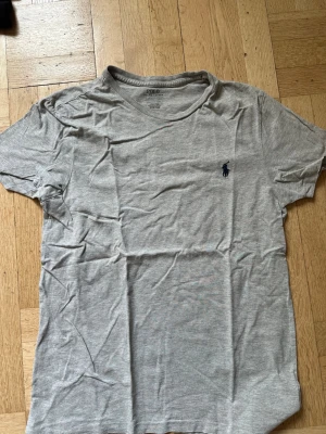 Grå t-shirt från Polo Ralph Lauren - Snygg grå t-shirt från Polo Ralph Lauren med klassisk passform och korta ärmar. T-shirten har en liten mörkblå logga broderad på bröstet och är tillverkad i mjuk bomull. Perfekt basplagg till garderoben.