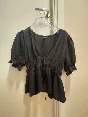 Svart blus med puffärm från Gina Tricot - Trendig svart blus från Gina Tricot med puffiga korta ärmar och v-ringning. Blusen har en smockad struktur, knytband i midjan och volang nedtill. Perfekt för dig som gillar en feminin och avslappnad stil.