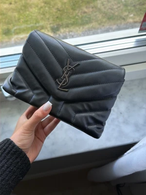 Axelväska i skinn - Säljer en axelväska med klassisk logga i metall framtill. Väskan är quiltad i mjukt skinn och har en stilren, modern design med vikbar öppning. Perfekt accessoar för att lyfta vilken outfit som helst. Kommer med band.