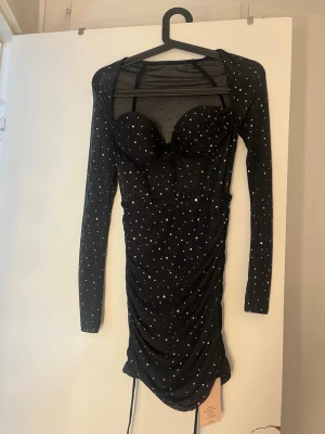 Svart glittrig kort klänning - Supersnygg svart kort klänning med långa ärmar och glittriga detaljer. Klänningen har en figurnära passform, mesh-tyg upptill och vadderade kupor. Perfekt för dig som vill sticka ut med en trendig look.