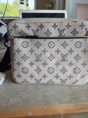 Louis Vuitton beige handväska monogram - Beige handväska från Louis Vuitton med ikoniskt monogrammönster i svart. Väskan har en rektangulär form, ytterfack med dragkedja och justerbar axelrem. Tillverkad i tålig canvas med svart textilfoder. Perfekt accessoar för dig som gillar exklusiv design.