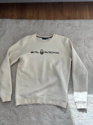 Vit sweatshirt från Sail Racing - Säljer en stilren vit sweatshirt från Sail Racing med svart logotyptryck på bröstet. Tröjan har rund halsringning, långa ärmar och ribbade muddar vid ärmslut och nederkant. Perfekt för en clean och sportig look.
